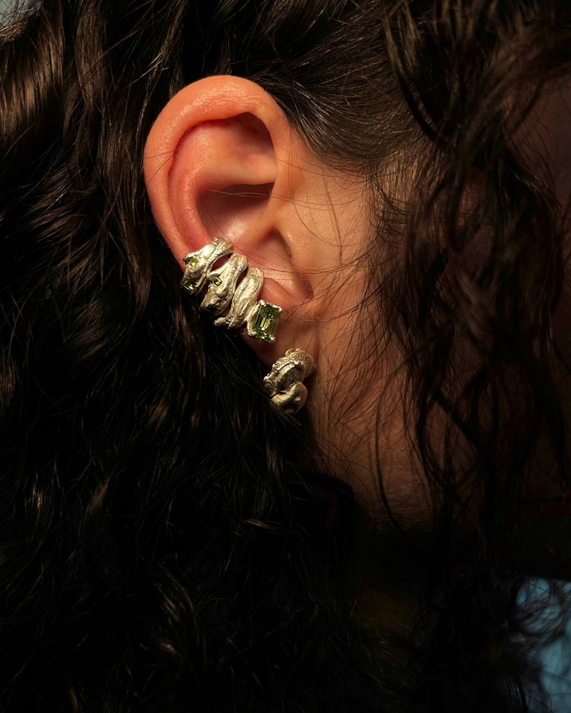 Close-up of an ear wearing a gold earring with green gemstones, surrounded by dark hair.